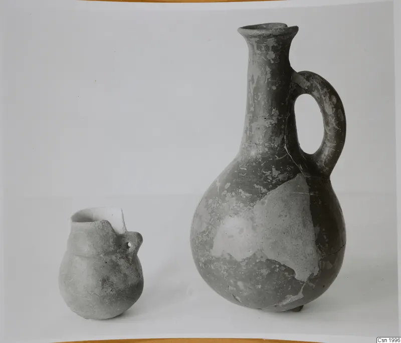 Two pottery jugs on a pale surface: a tall jug with a loop handle on the right and a small handled jug on the left.