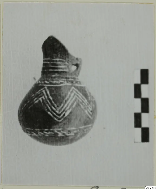 Pottery vessel with a narrow neck, side loop handle and light chevron bands, beside a checkered scale bar.