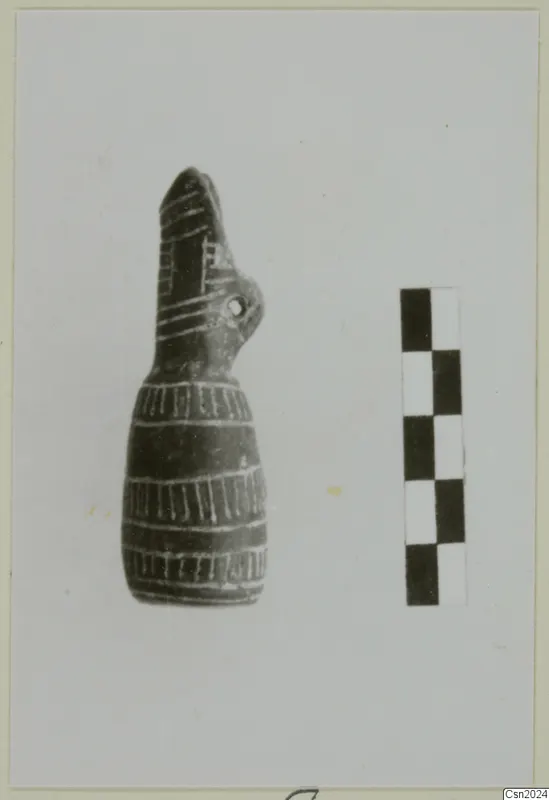 Dark pottery vessel with a tall beak-like top and pale banded lines, photographed beside a scale bar