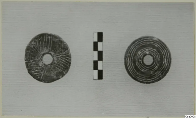 Two flat discs with central holes, one with radiating incised lines and one with concentric rings, beside a checkered scale bar