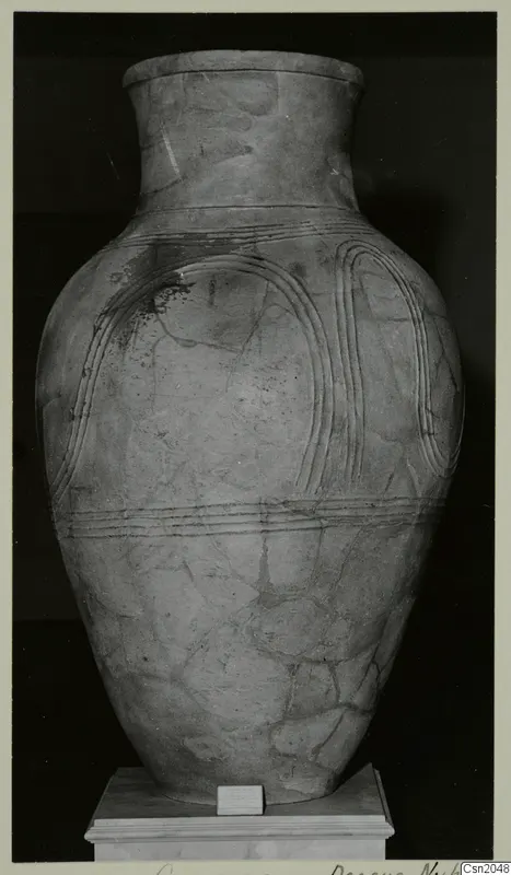 Large pottery pithos with a tall neck and incised ridged bands, standing on a plinth in a museum display.