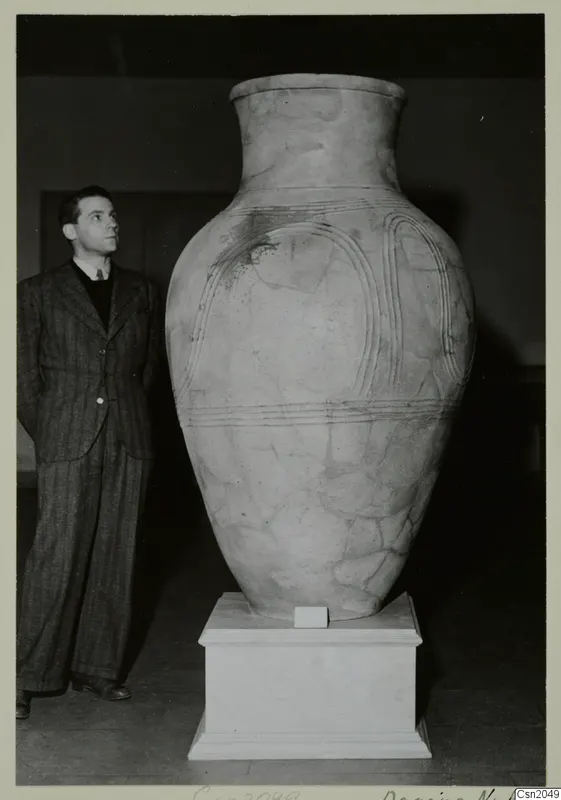 Large pale pottery vessel on a white pedestal, with a man in a striped suit standing beside it and looking up at it.