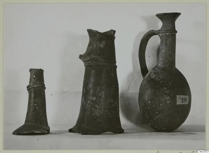 Three pottery vessels of Base-Ring Ware on a pale surface: two broken pieces and one complete juglet with a single handle.