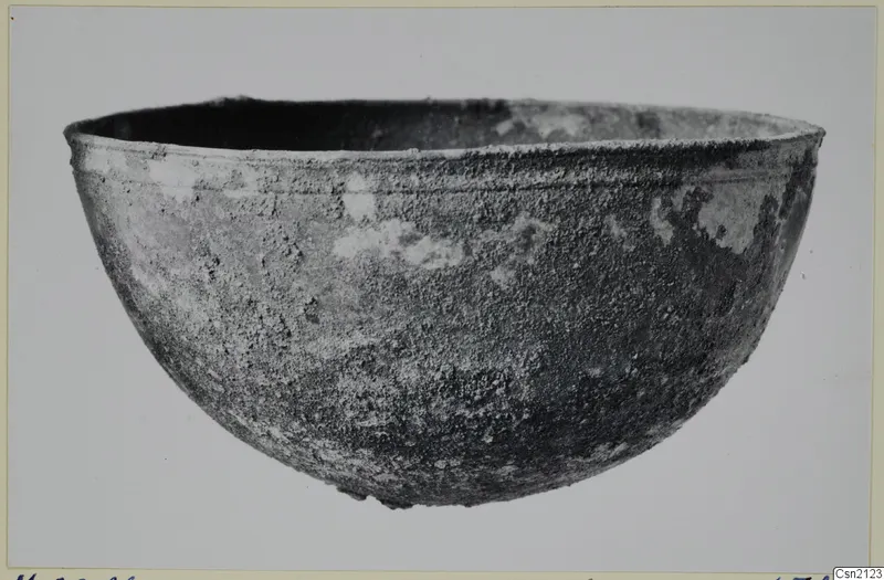 Rounded bowl with a flared rim and an encrusted, corroded surface, shown in side view on a plain background.