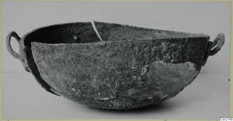 Corroded bowl with two loop handles, seen from the side against a plain background.