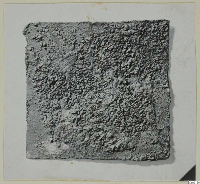 Corroded metal fragment, a flat square piece with a crusted grey surface, photographed on a card mount