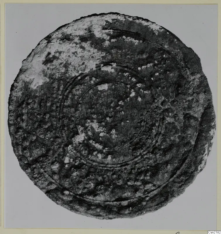 Corroded bronze disc with raised concentric rings and a pitted, bumpy surface, photographed on a grey backdrop.