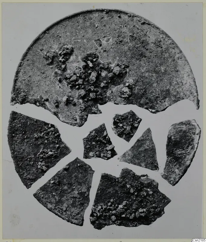 Corroded bronze lid with several broken fragments arranged around it, photographed on a pale grey background
