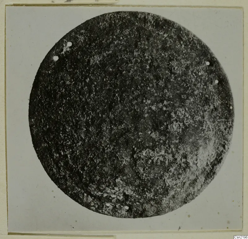 Round metal disc fragment with small rivet holes around its edge, photographed on a card mount