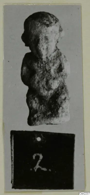 Small standing figure of a person with arms at the sides, photographed on grey card above a number label