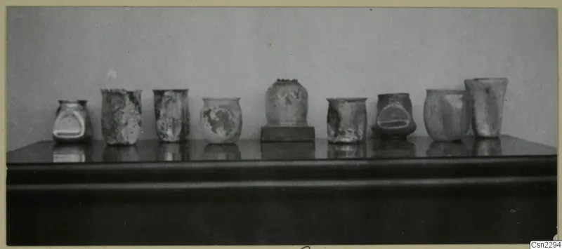 Row of weathered blown glass jugs and vases from Vouni, Korakas tombs, standing on a dark shelf.
