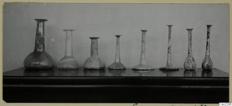 Row of several blown glass vessels with long slender necks standing on a dark shelf against a grey backdrop