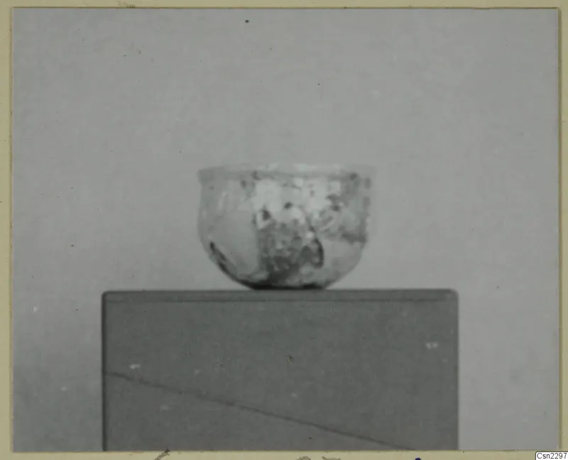 Small thin-walled vessel with a wide mouth, standing on a dark block against a grey backdrop