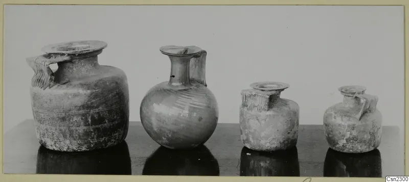 Four blown glass jugs and jars with handles standing in a row on a dark table.