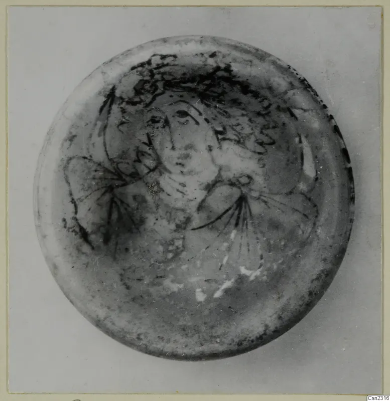 Round glass object with a dark painted face and hair pattern, photographed from above.