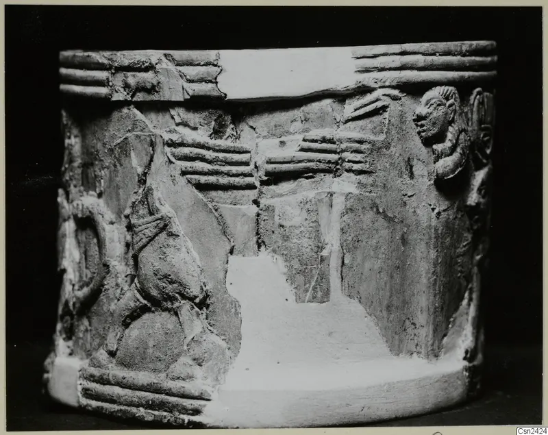 Cylindrical carved object with relief figures and ridged bands, photographed against a black background