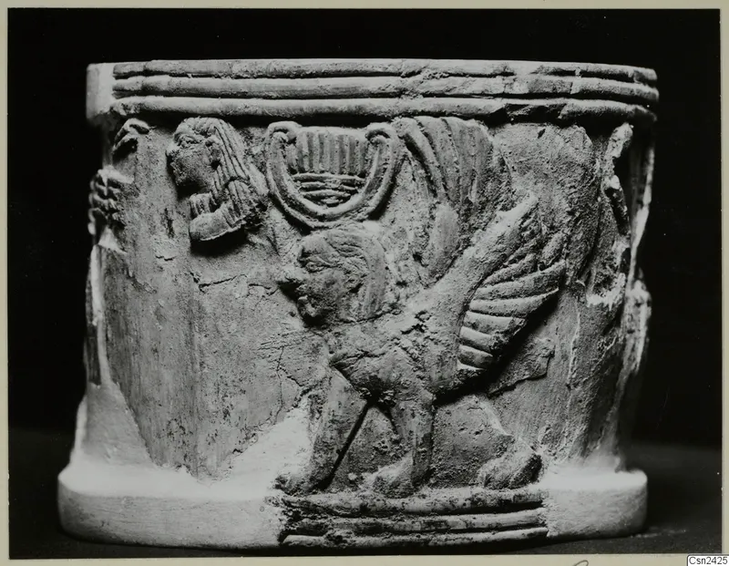 Carved cylindrical object with a relief of a winged creature and a seated figure, shown against a dark background