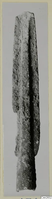 Corroded metal weapon blade with a central ridge, photographed upright against a grey background.