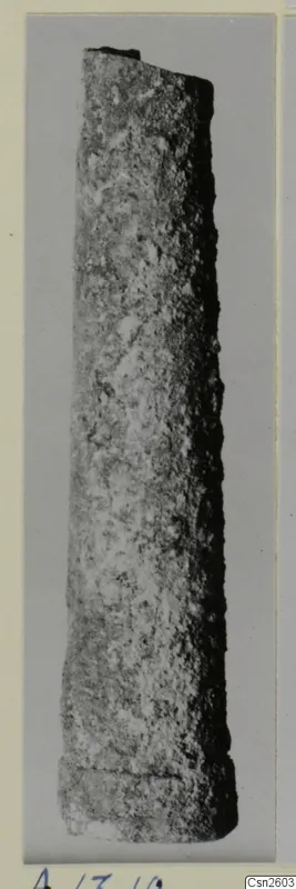 Long, tapering bronze object with a corroded surface, probably a weapon fragment, shown upright against a grey background
