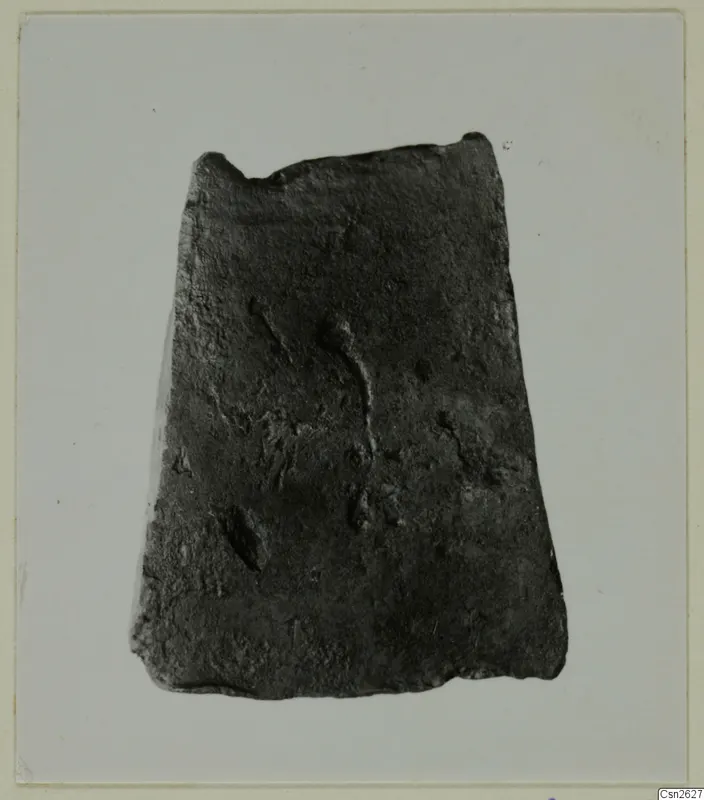 Corroded flat metal object with a broken top edge, photographed on a pale grey backdrop