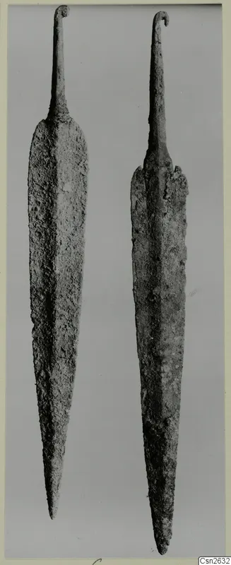 Two corroded metal weapons with long tapering blades and hooked tangs, shown side by side against a grey background.