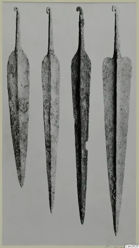 Four long metal weapon blades standing upright side by side, each with a curled tang at the top, on a grey background.
