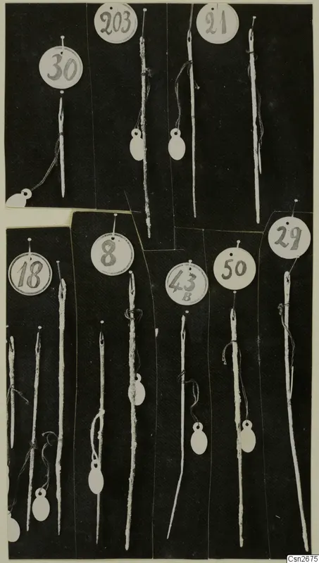 Bronze pins or needles, each tied to a round numbered paper tag, laid out on a dark background