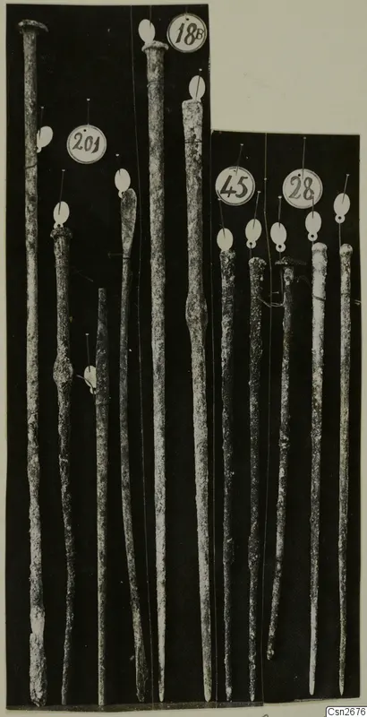 Long slender bronze tools with tapering shafts, standing upright against a black background with numbered paper tags.