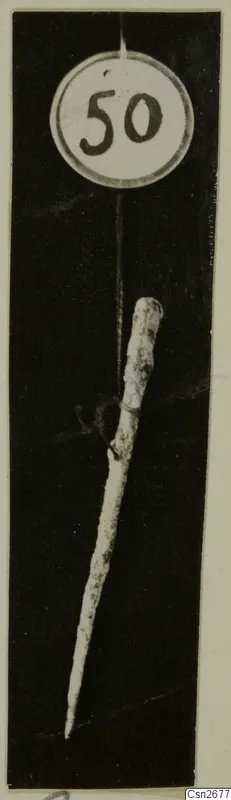 Slender metal tool with a pointed tip, hung by a thread from a round number label against a black background