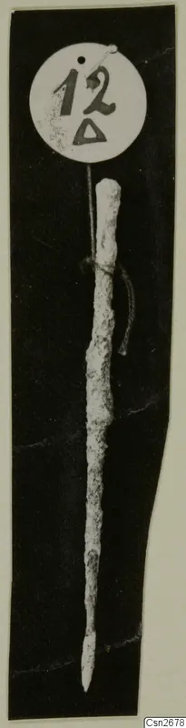 Corroded bronze implement with a pointed tip, tied with string to a round number label, photographed on black.