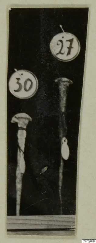 Two bronze tools with flared tops and long tapering shafts, each beside a round number label, against a dark background.