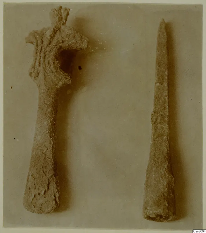 Two corroded bronze tools on a plain background: a larger one with a flared, curved upper end and a shorter, long pointed one beside it.
