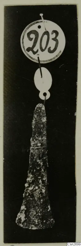 Corroded metal tool hanging from two white tags against a dark background