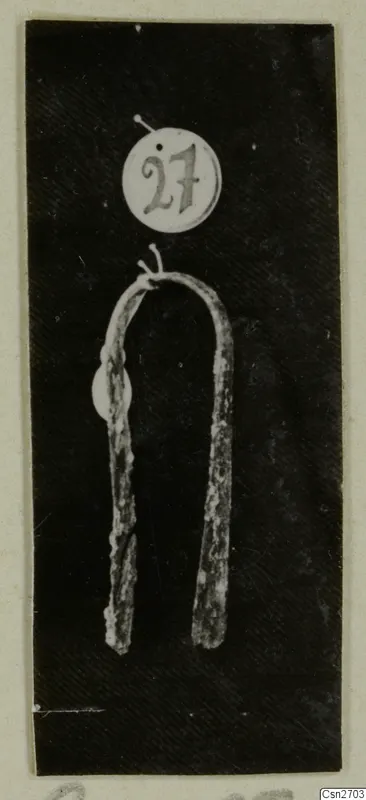 Bronze tool with two long arms joined by a curved bend, shown with a round number label, on a dark background.