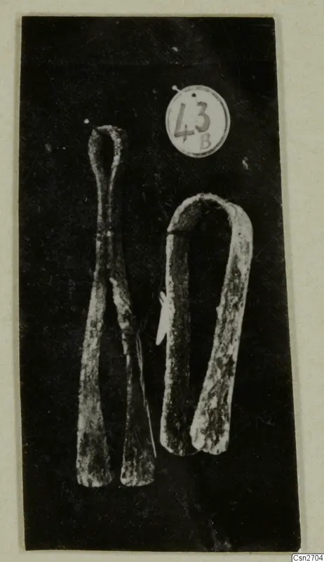 Two corroded bronze tools on a dark background, one a pair of tongs with a looped top and flat ends.