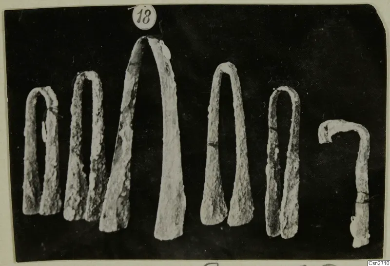 A row of corroded bronze tools with tapering arched arms against a dark background.