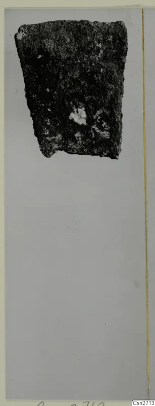 Corroded dark metal fragment, possibly a tool, photographed on a grey card mount.