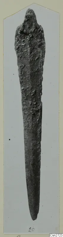 Long corroded bronze tool with a tapering blade and rounded upper end, shown on a grey card