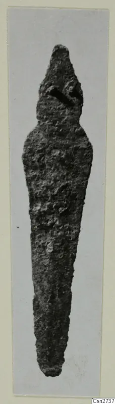 Long, tapering corroded metal object standing upright, photographed on a grey mount
