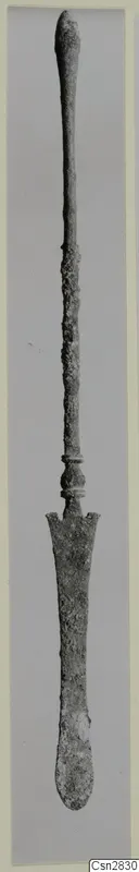Long bronze implement with a slender shaft and a flat spatula-like blade at the lower end, photographed upright on a grey backdrop.