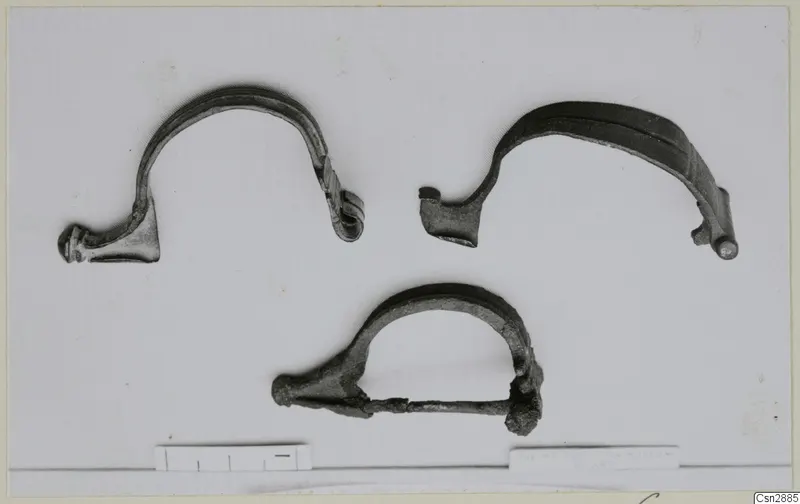 Three bronze bow-shaped personal adornments on a pale background, with a scale bar along the bottom edge.