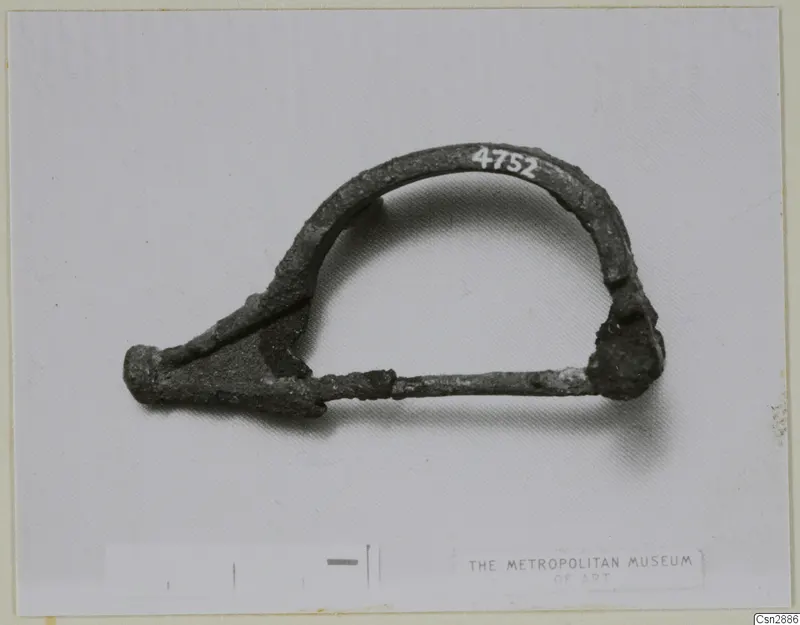 Bronze bow-shaped brooch with a long pin, photographed flat beside a scale bar