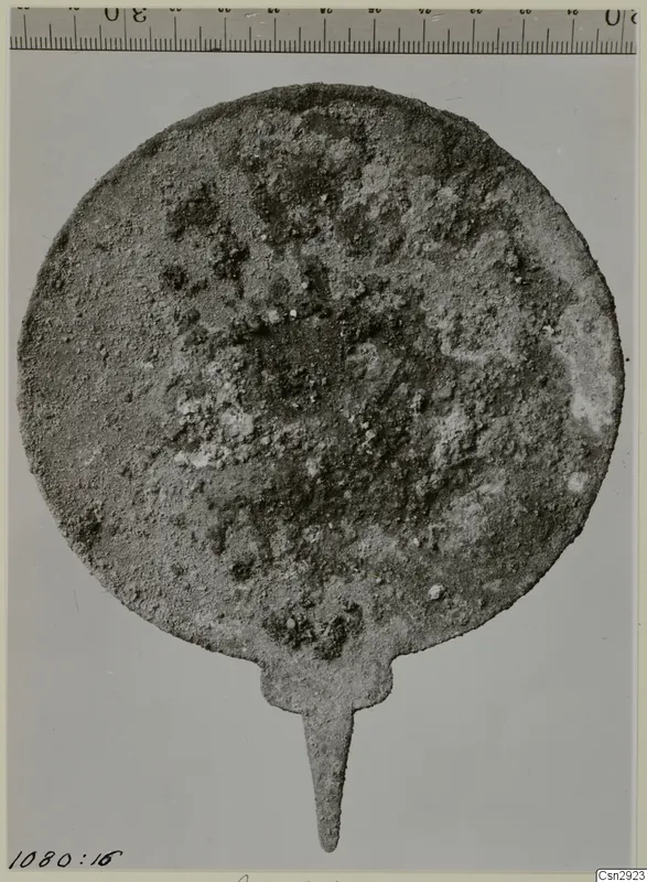 Corroded round metal disc with a pointed tang, photographed flat with a ruler along the top edge