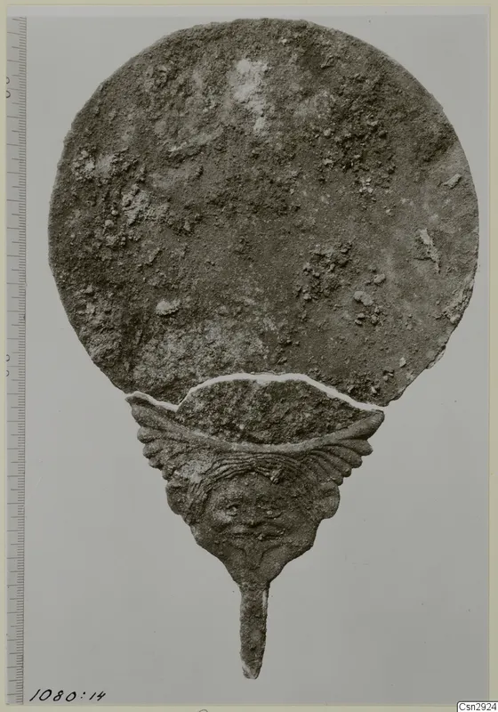Corroded metal disc with a handle ending in a human face, photographed beside a scale bar