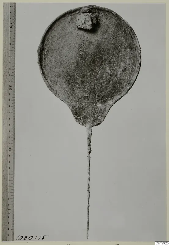 Corroded round metal disc with a long thin stem, shown beside a measuring rule.