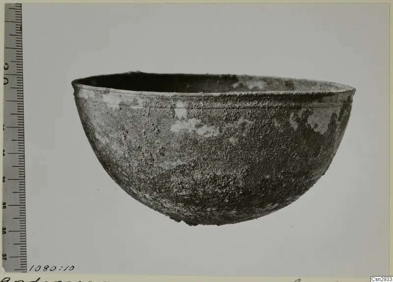 Corroded bowl with a rounded body and a thick rim, photographed beside a scale ruler