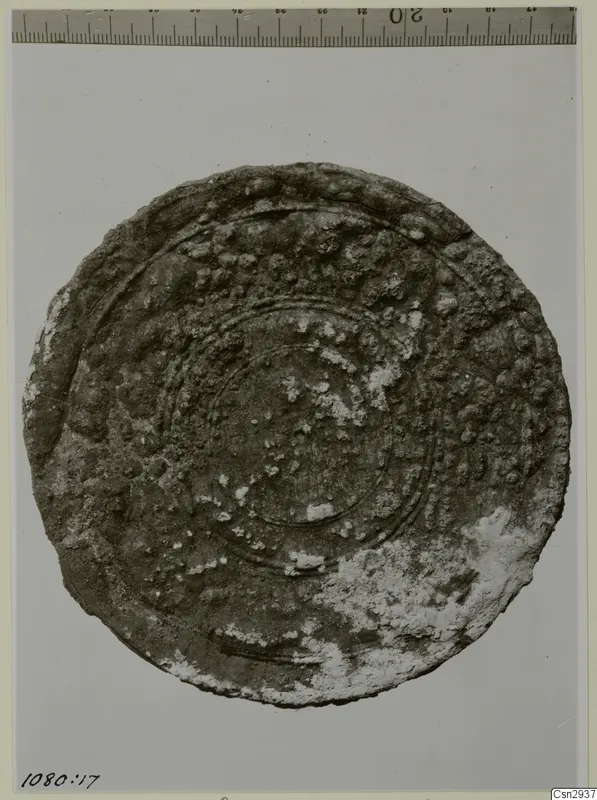 Round, heavily corroded object with concentric ridges and a pale crust, shown against a grey background with a scale above.