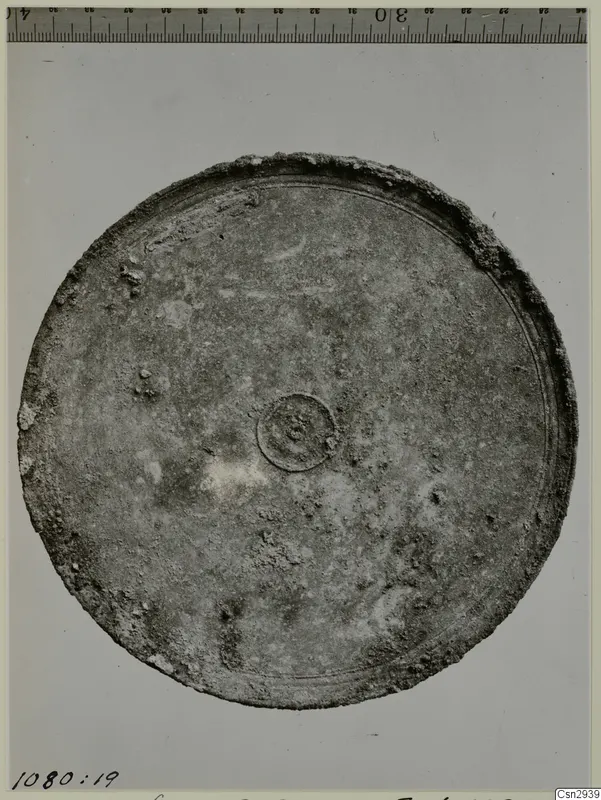 Round metal object with a raised rim and small central boss, photographed beside a scale ruler