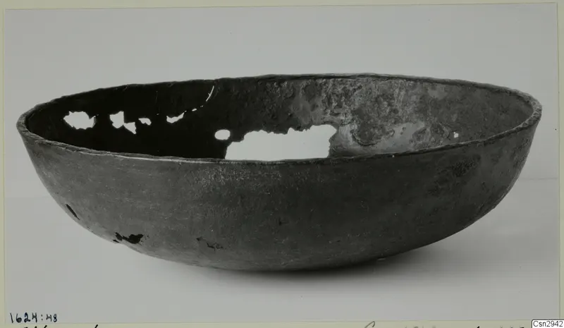 Dark grey bowl with a rounded body and rim, with holes broken through its wall, photographed on a plain background.