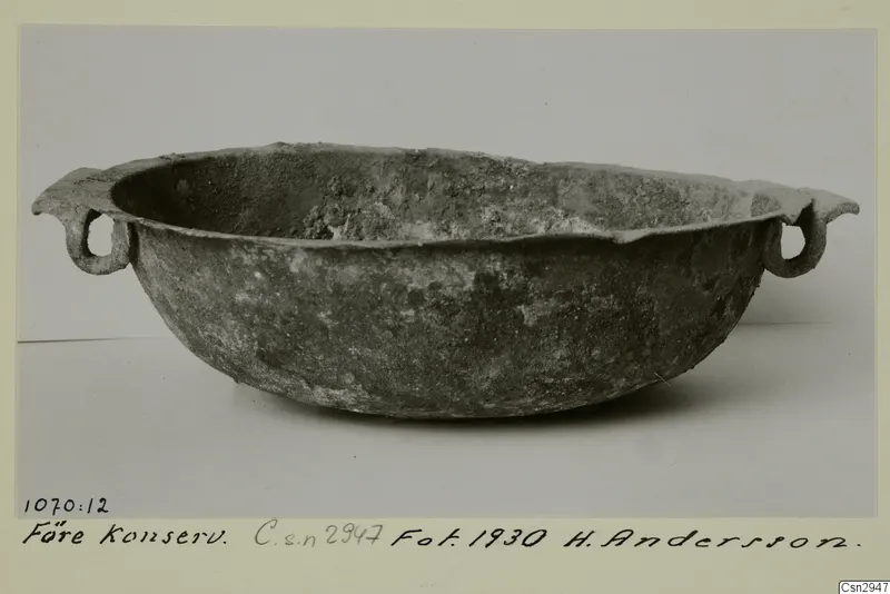 Corroded metal bowl with two loop handles, photographed before conservation